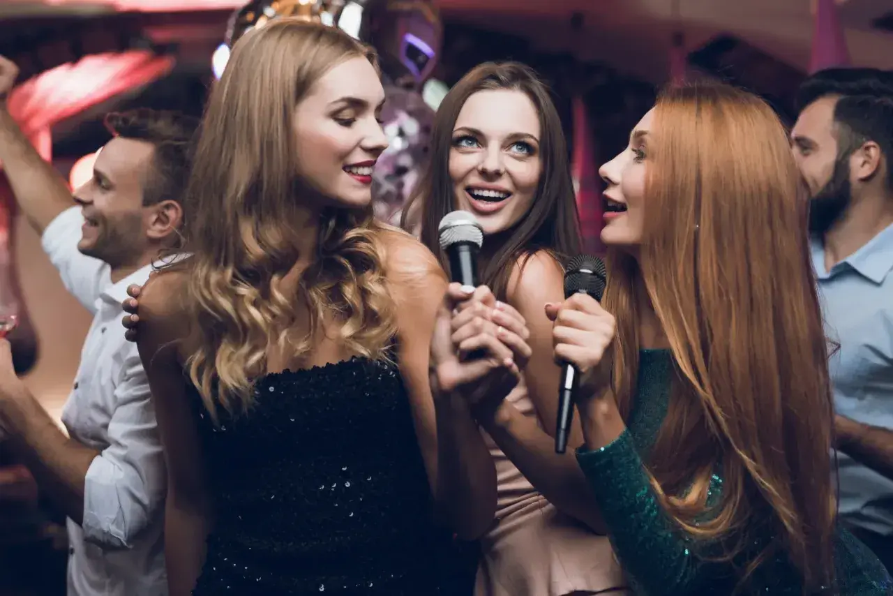 Trzy kobiety śpiewają do mikrofonów, bawiąc się przy najlepszych karaoke. W tle widać innych ludzi na imprezie.
