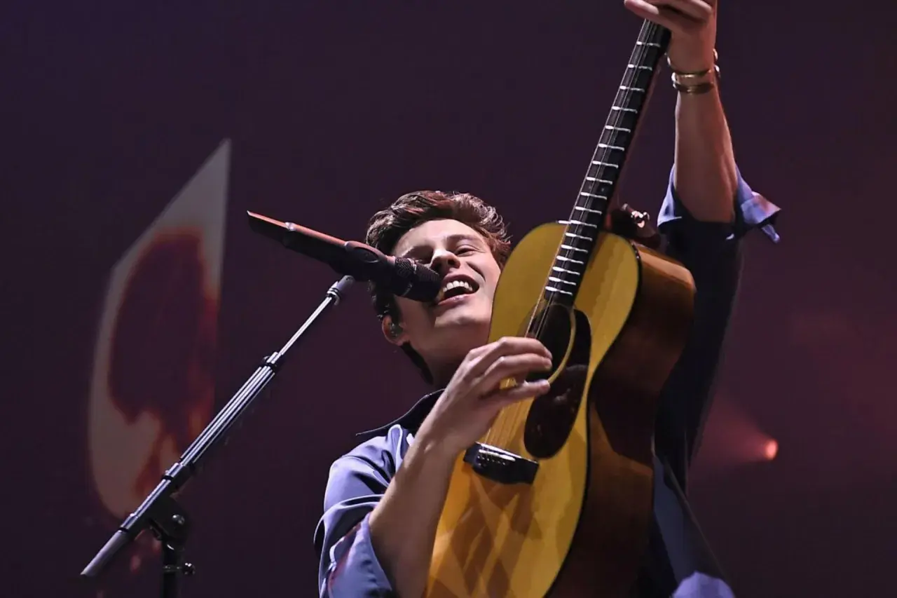 Shawn Mendes na koncercie, śpiewa i gra na gitarze, wznosząc ją w górę.