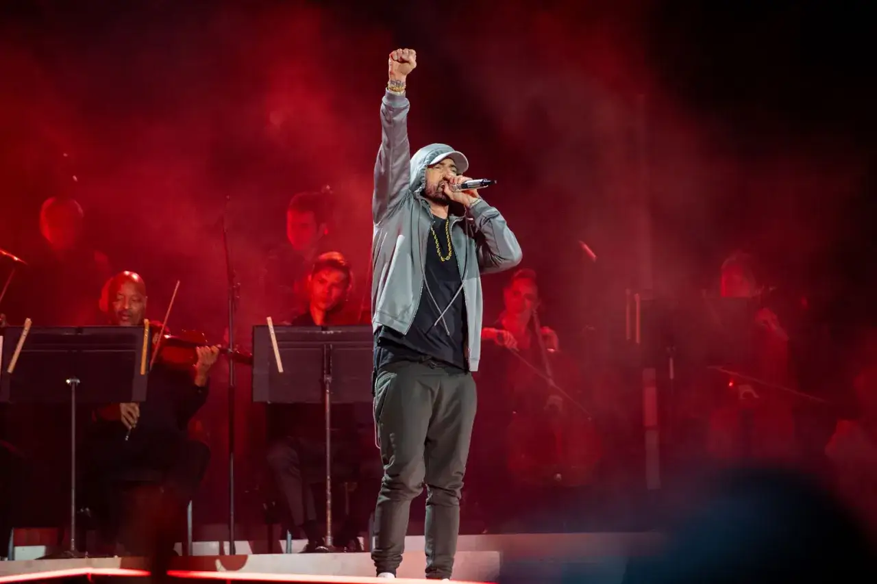 Eminem na scenie z orkiestrą, wykonuje swoje utwory. Wzniesiona pięść i mikrofon w dłoni.