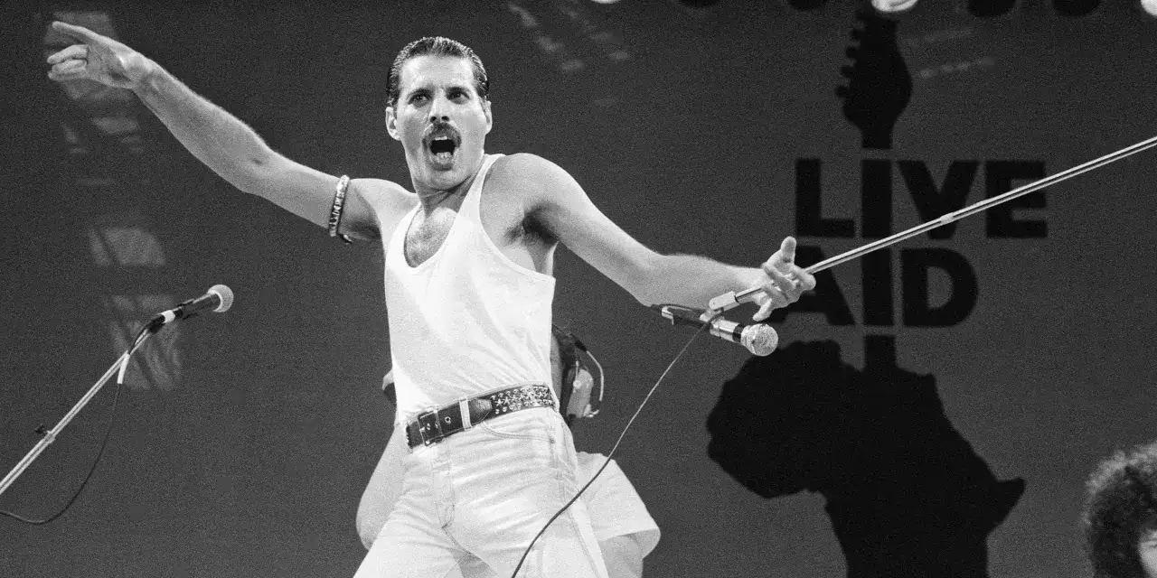 Freddie Mercury w białej koszulce i dżinsach, śpiewający na scenie Live Aid, z charakterystycznym wąsem i gestem ręki.