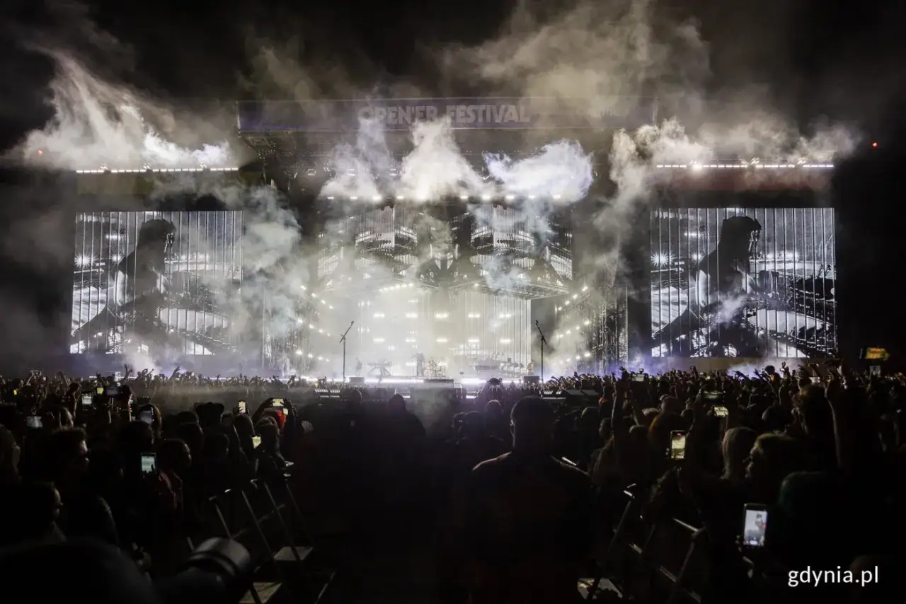Koncert Linkin Park Polska na Open'er Festival. Tłum ludzi z telefonami, dym i światła na scenie.