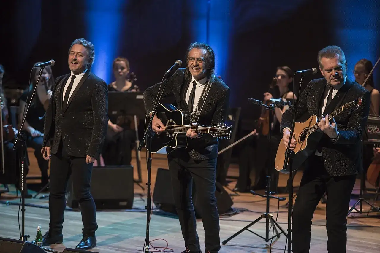 Trzech mężczyzn w błyszczących marynarkach śpiewa i gra na gitarach. W tle orkiestra. To vox piosenki alfabetycznie.