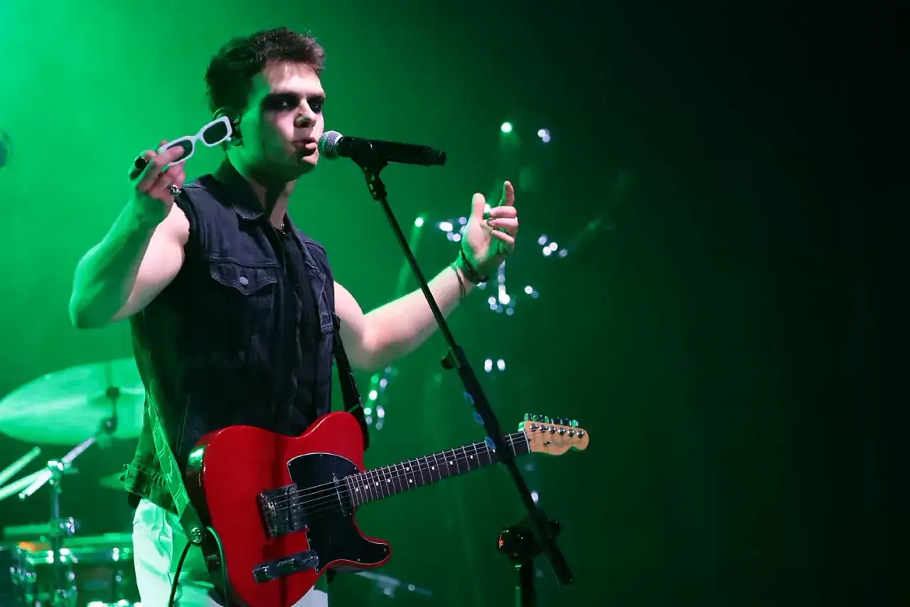 Kris Cugowski w czarnej kamizelce i z czerwoną gitarą na scenie, w zielonym świetle.