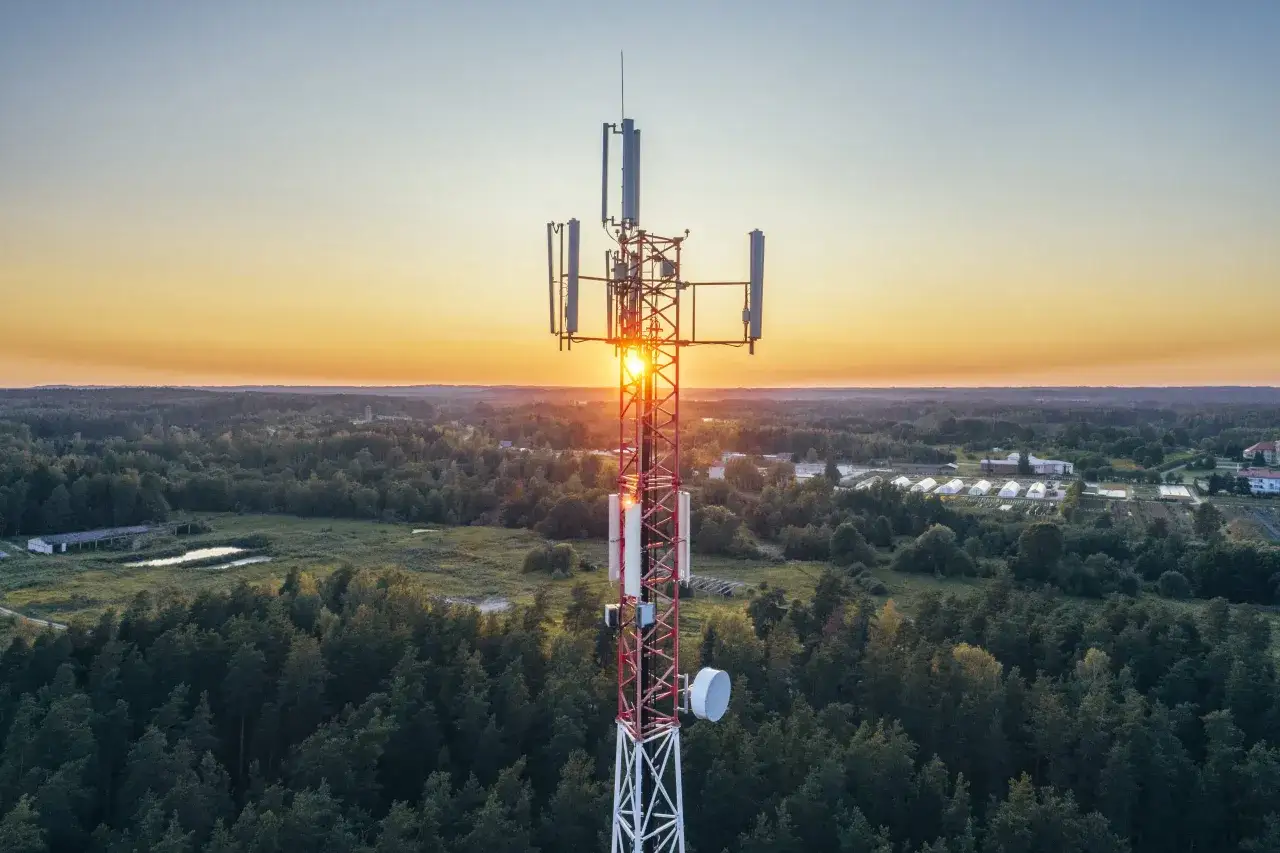 Wieżowa radiostacja na tle zachodzącego słońca, otoczona lasem i polami.