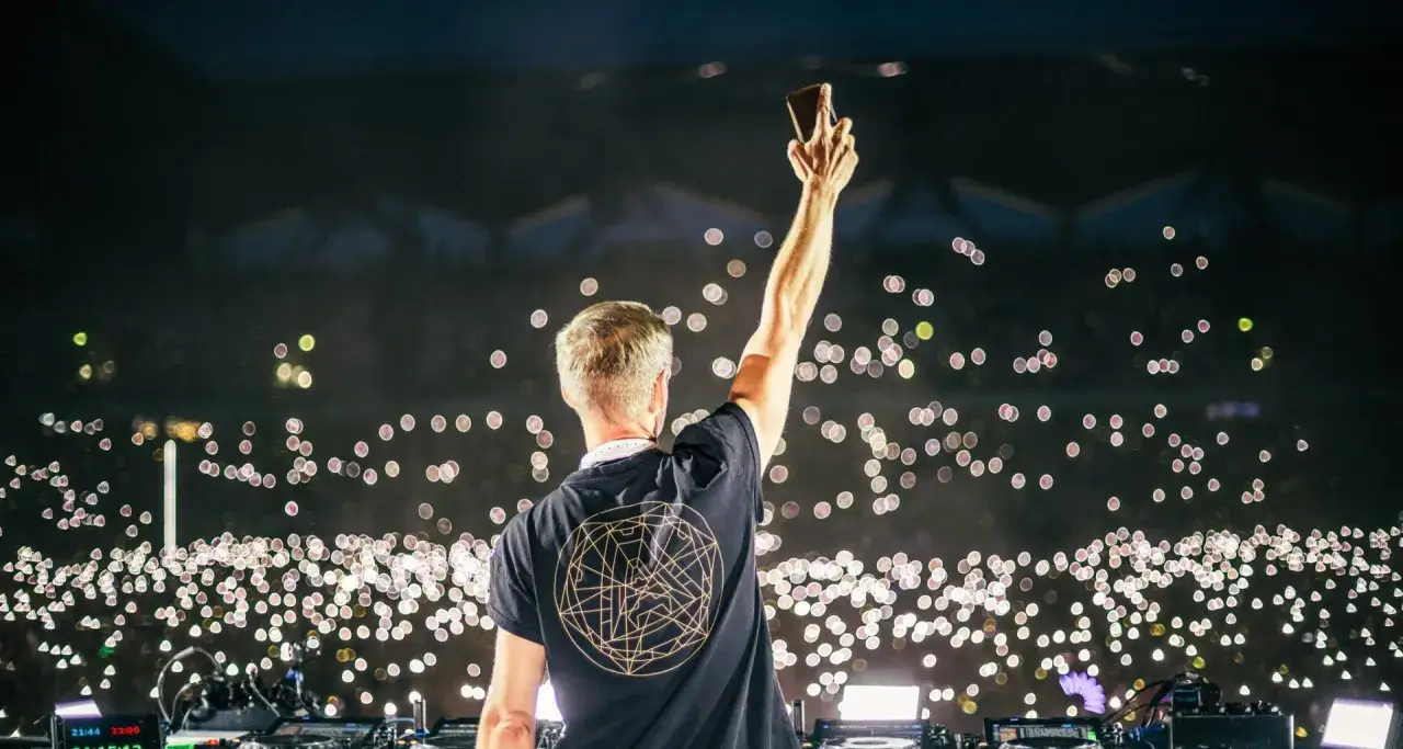Armin van Buuren w Warszawie, DJ z ręką w górze pozdrawia tłum rozświetlony tysiącami świateł telefonów.