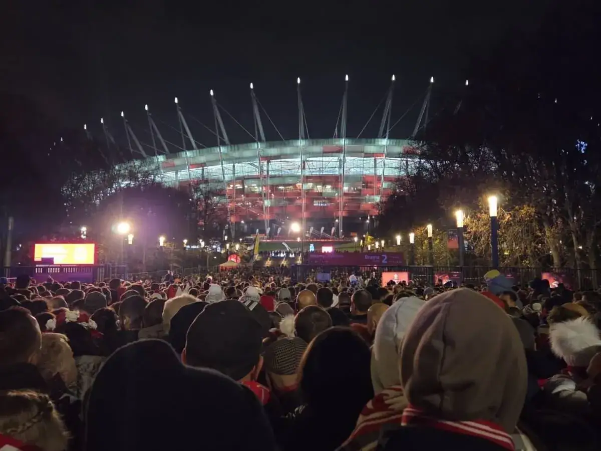 Tłum ludzi przed roztańczonym PGE Narodowym nocą. Stadion świeci na czerwono, a nad głowami widać lampy i drzewa.