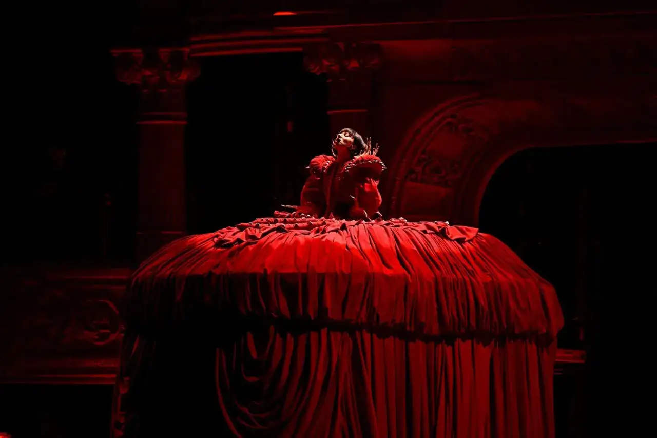 Lady Gaga w czerwonej, obszernej sukni na scenie podczas koncertu. Artystka w teatralnej pozie.