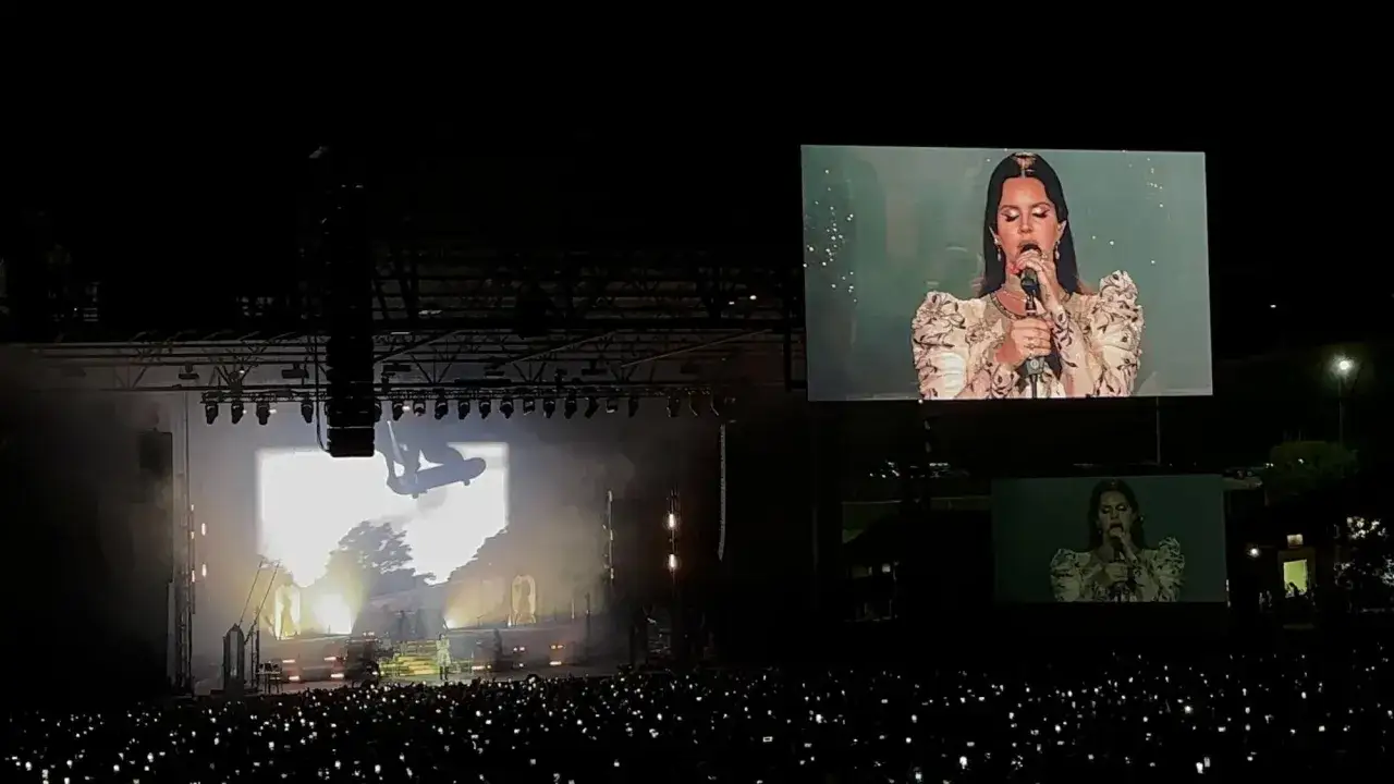 Lana Del Rey na koncercie, tłum z telefonami, na ekranie artystka w sukni z bufiastymi rękawami.