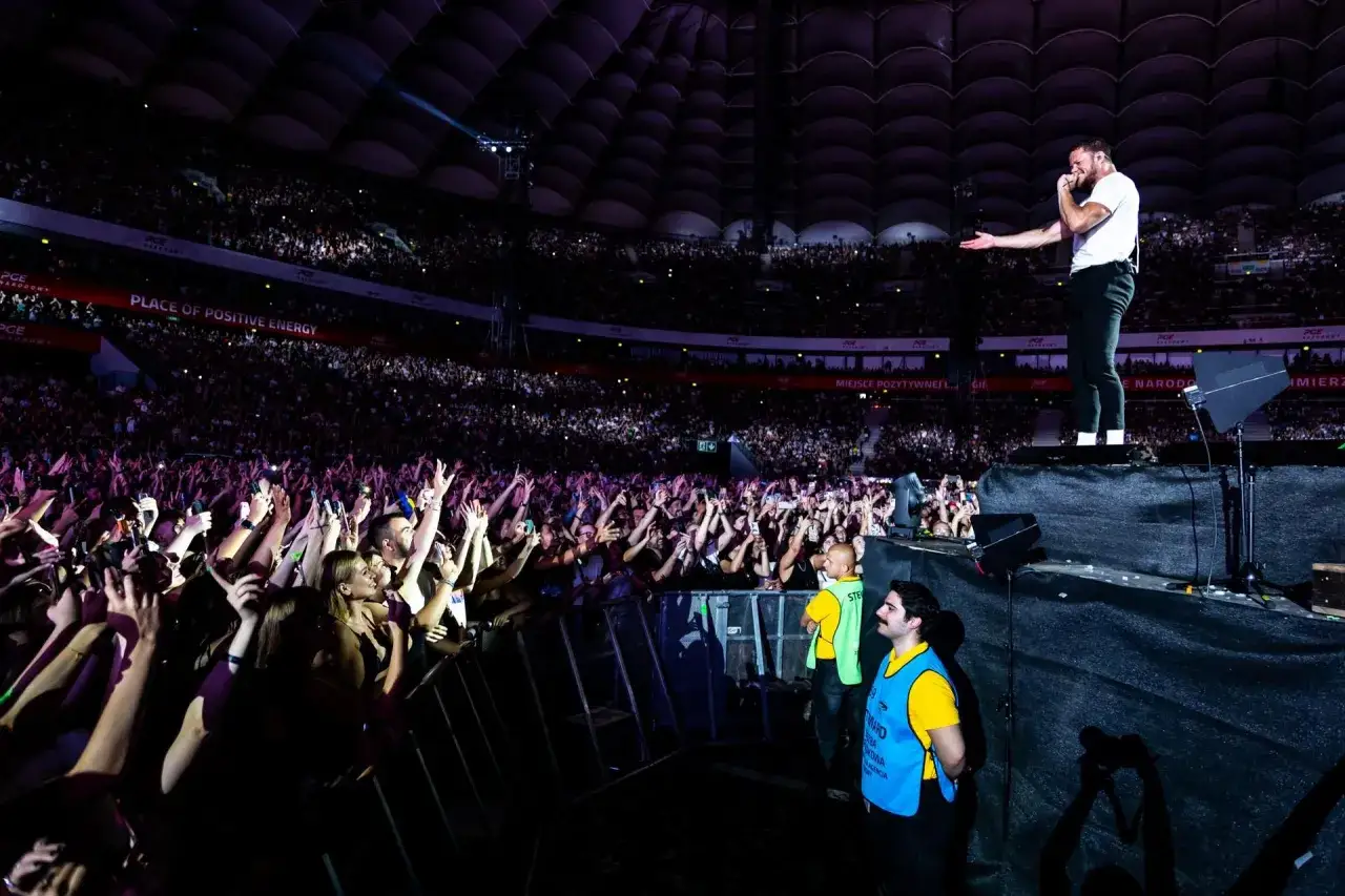 Imagine Dragons koncert na PGE Narodowym. Tłum fanów z telefonami w górze, wokalista na scenie, światła i energia.