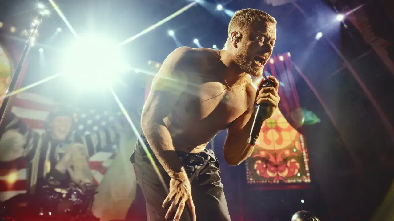 Imagine Dragons koncert Polska: wokalista z odsłoniętym torsem śpiewa do mikrofonu na tle amerykańskiej flagi i scenografii.