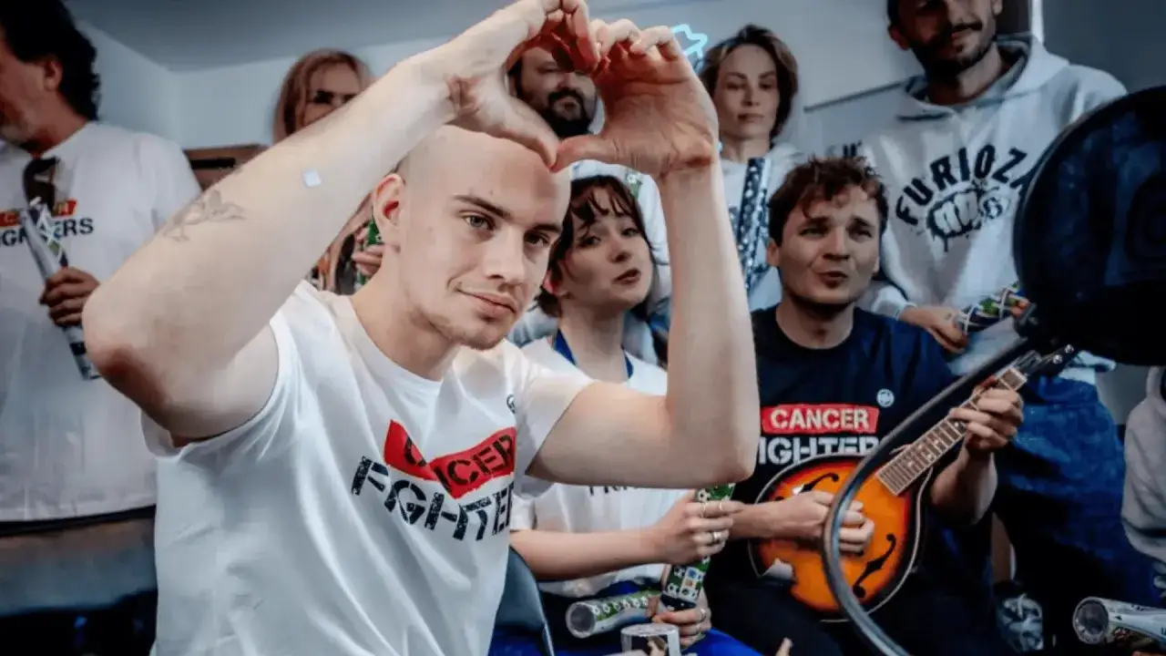 Grupa ludzi podczas łatwogang stream. Mężczyzna z przodu robi serce dłońmi. W tle grający na mandolinie.