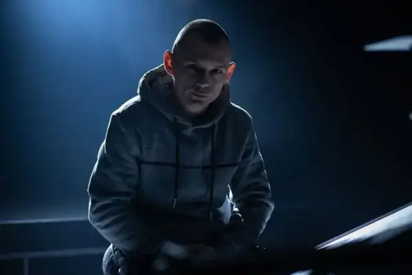 Młody raper w szarej bluzie z kapturem, oświetlony od tyłu, wpatruje się w obiektyw.